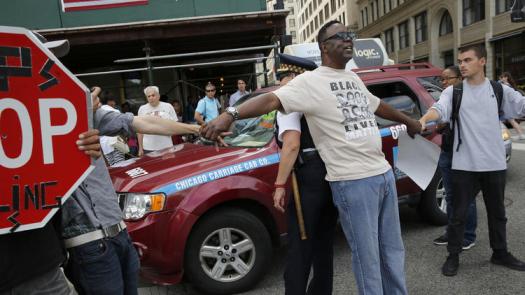A photo of a black man wearing a "black lives matter" shirt linking arms with two other protestors. His back is to a police officer and there is a red car behind them.